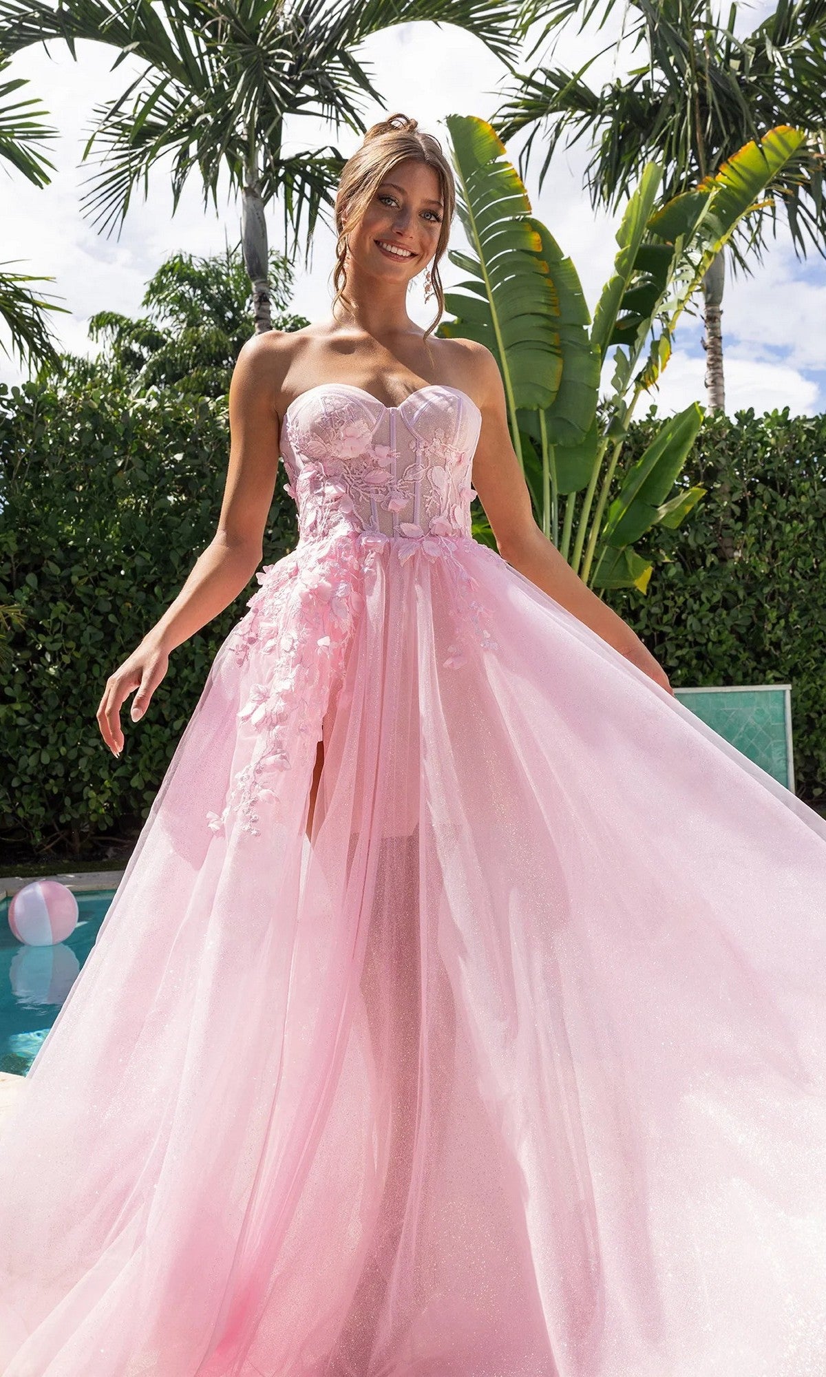A woman in a pink Portia and Scarlett PS26078 long formal dress with a corset bodice and glitter tulle skirt stands by a pool, surrounded by palm trees and lush greenery.